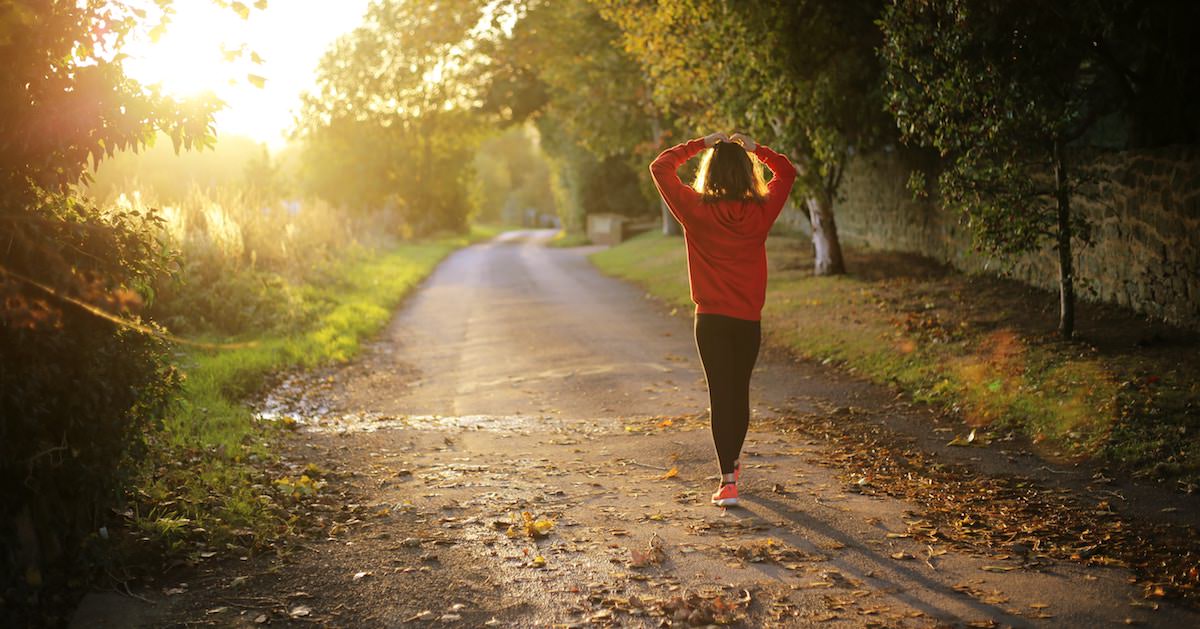 A woman walking down a road with your hands crossed over her head in a stretch. It is a fall morning. Brown leaves outline the rode, and the sunrises in the east. It is part of her morning routine.