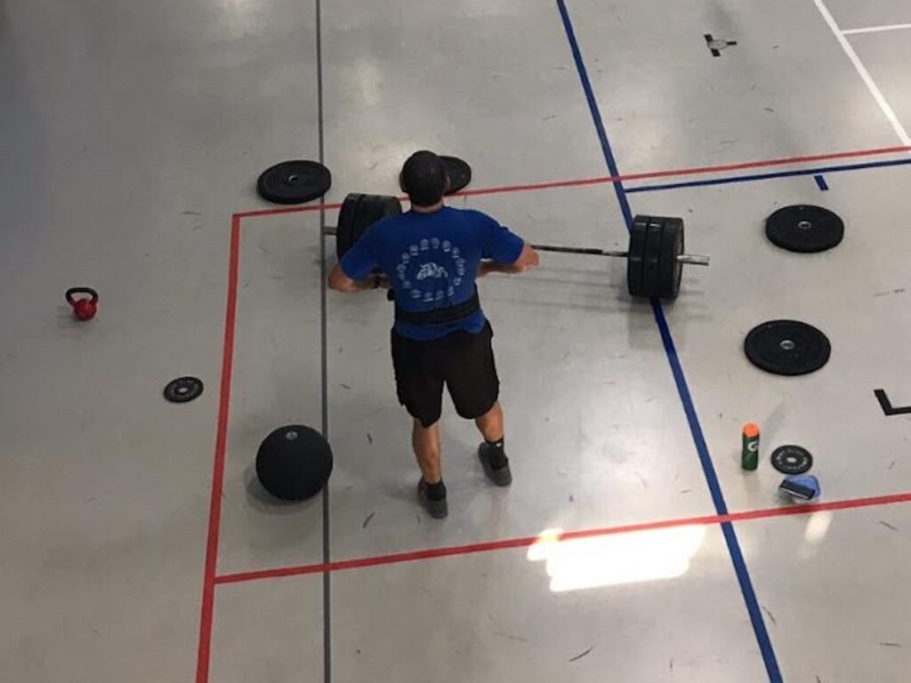 Man in a blue shirt demonstrating consistency over intensity by lifting a barbell in a gym, embodying the mantra of regular effort post-injury