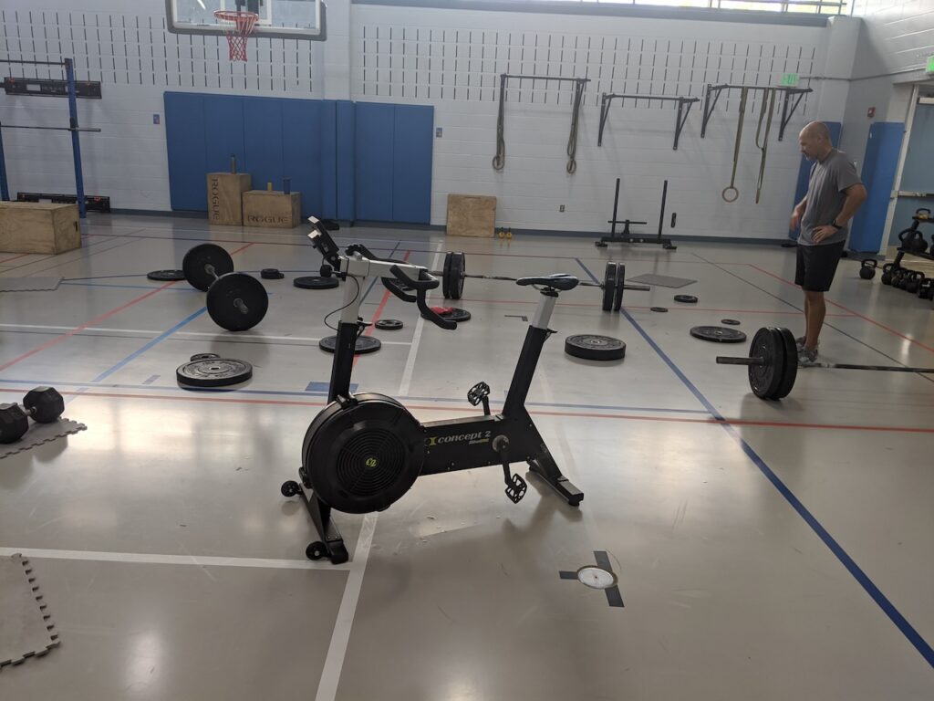 Man in a gym showcasing the benefits of functional fitness with diverse equipment like barbells, dumbbells, and a Concept2 cycling machine.