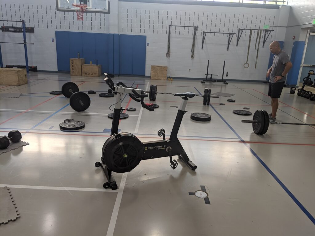 Man in a gym showcasing the benefits of functional fitness with diverse equipment like barbells, dumbbells, and a Concept2 cycling machine.