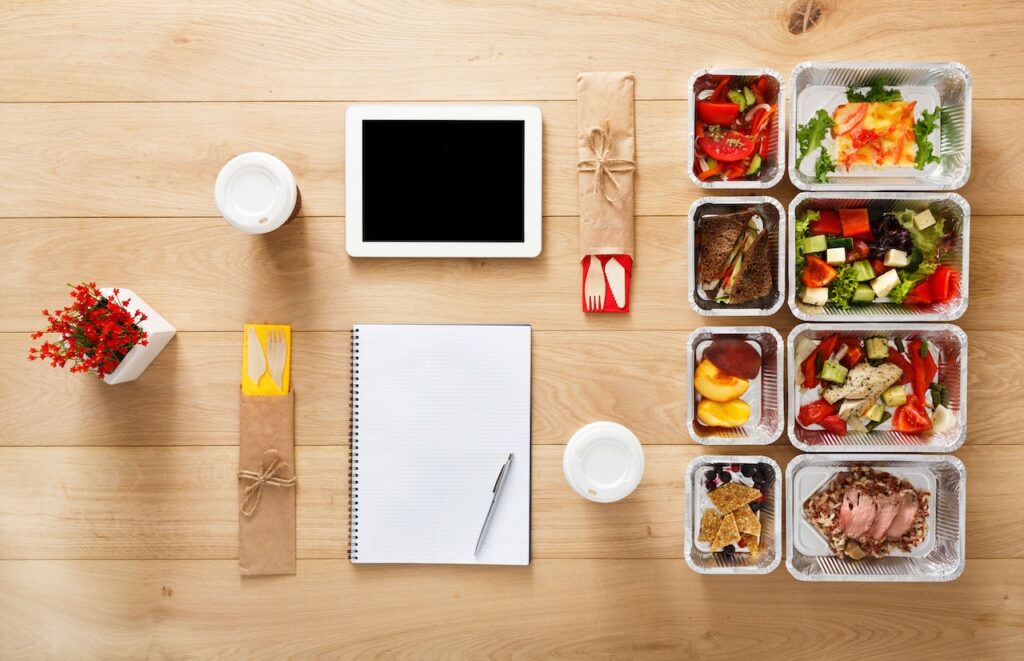 Healthy meal planning essentials with prepared meals, a digital tablet, notepad, and utensils on a wooden table.