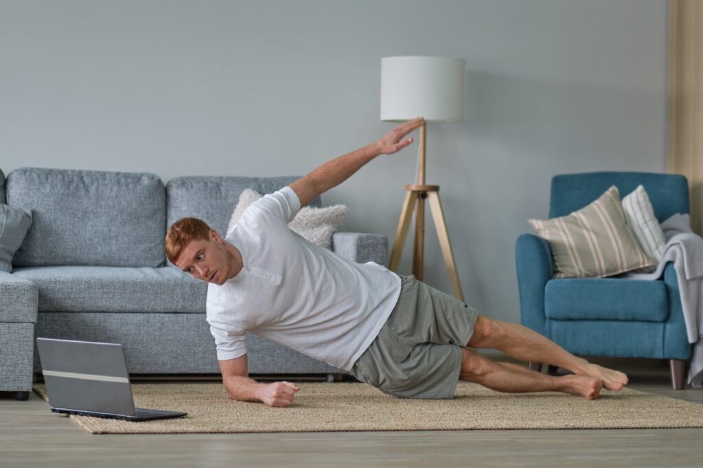 Man demonstrating a beginner-friendly side plank exercise while following a "workout for beginners" tutorial on his laptop.