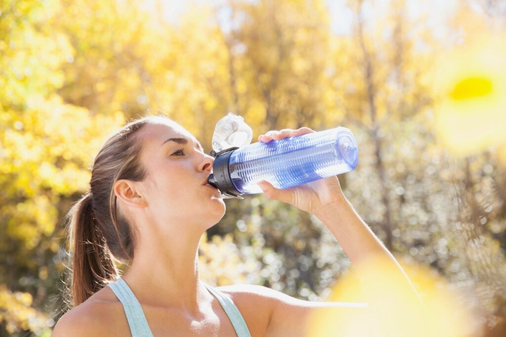 A woman exemplifying the recommendation for 'How much water should you drink' by sipping from a blue water bottle outdoors on a sunny day.