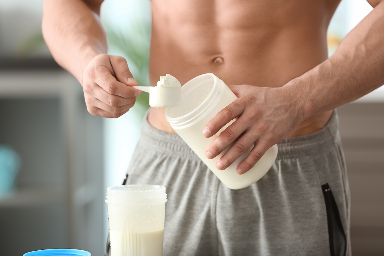 A person with a toned abdomen is scooping whey protein powder into a shaker bottle, preparing a supplement shake for muscle growth.