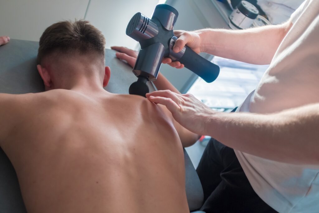 A person lying face down while receiving a massage with a handheld percussion therapy device during recovery days to aid muscle relaxation and recovery."