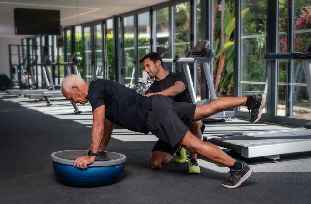 A senior man performing a plank exercise on a balance trainer with assistance from a personal trainer as part of a senior strength training program in a well-lit gym with exercise equipment in the background.