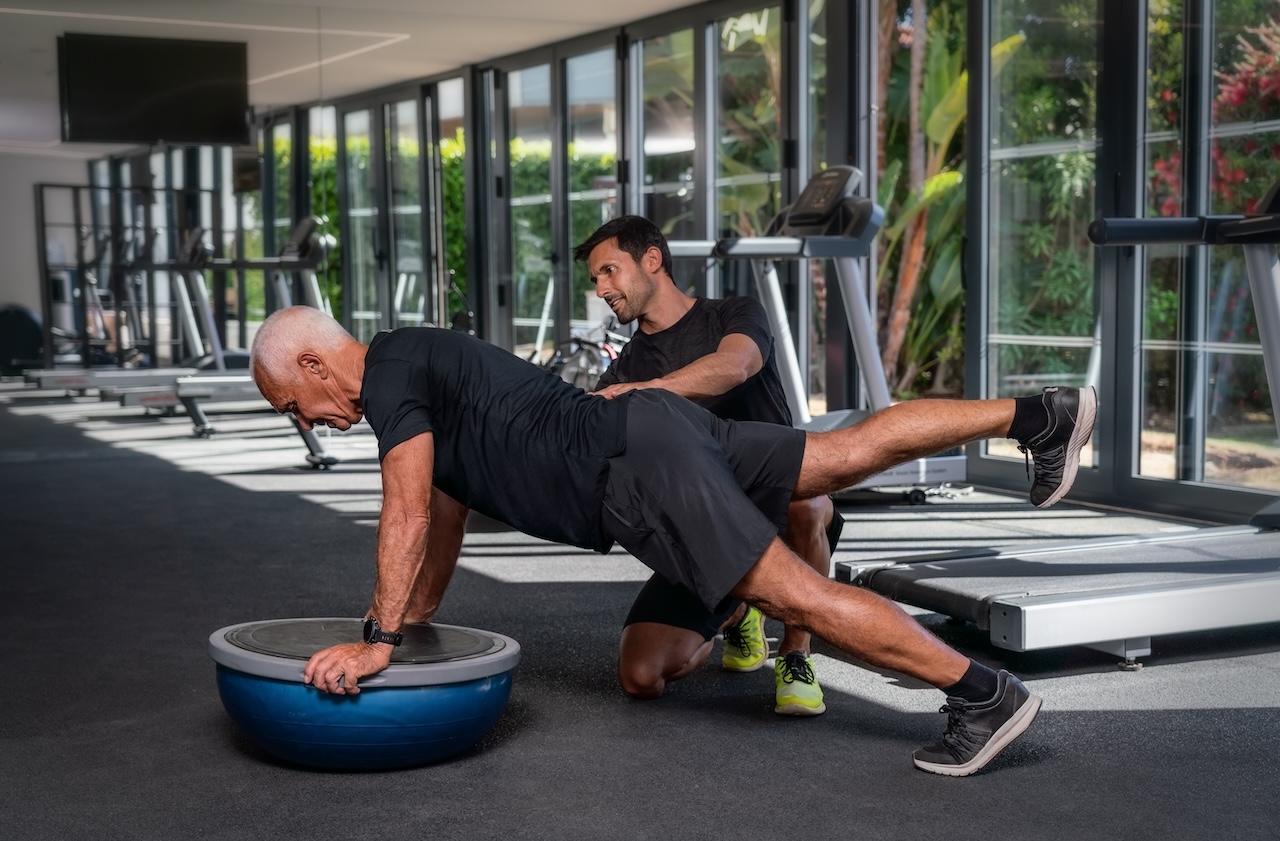A senior man performing a plank exercise on a balance trainer with assistance from a personal trainer as part of a senior strength training program in a well-lit gym with exercise equipment in the background.