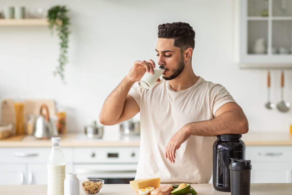 A man in a casual t-shirt is standing in a well-lit kitchen, drinking from a glass of milk. On the kitchen counter, there's a variety of high-protein foods including nuts in a bowl, sliced avocado, cheese, eggs, and slices of ham, arranged for preparing a meal. Next to him is a large black protein supplement jar and a shaker bottle, suggesting a focus on muscle gain nutrition.