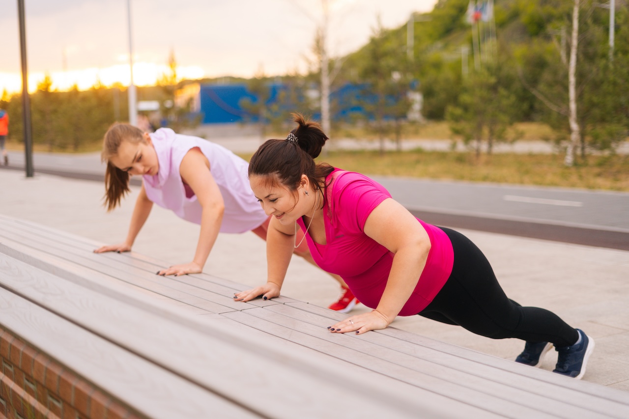 wo women engaging in strength training for weight loss outdoors by doing push-ups on a wooden bench in a park.