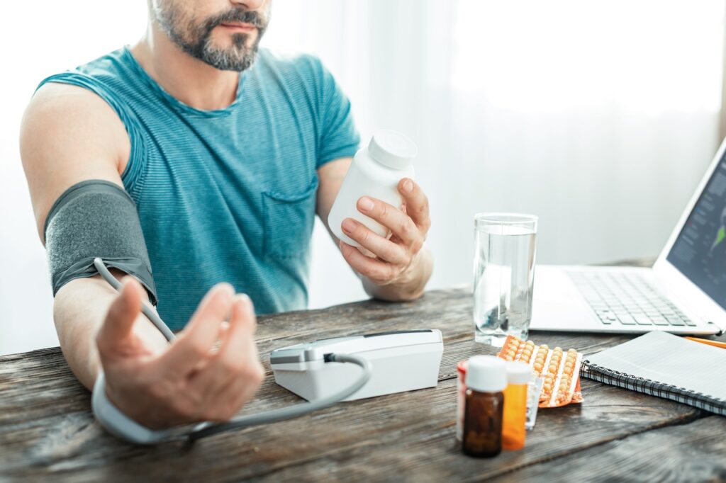 A man sits at a wooden table checking his blood pressure with a home monitor while holding a bottle of medication, with a glass of water, additional medication, and a laptop nearby, indicating a routine of health management potentially related to exercise with hypertension.