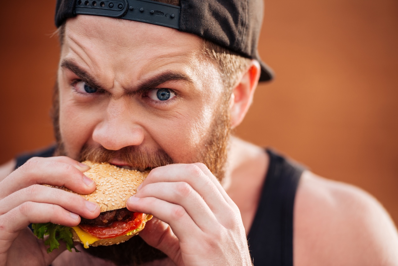 How to stop binge eating: A man pondering on the idea while voraciously biting into a cheeseburger.