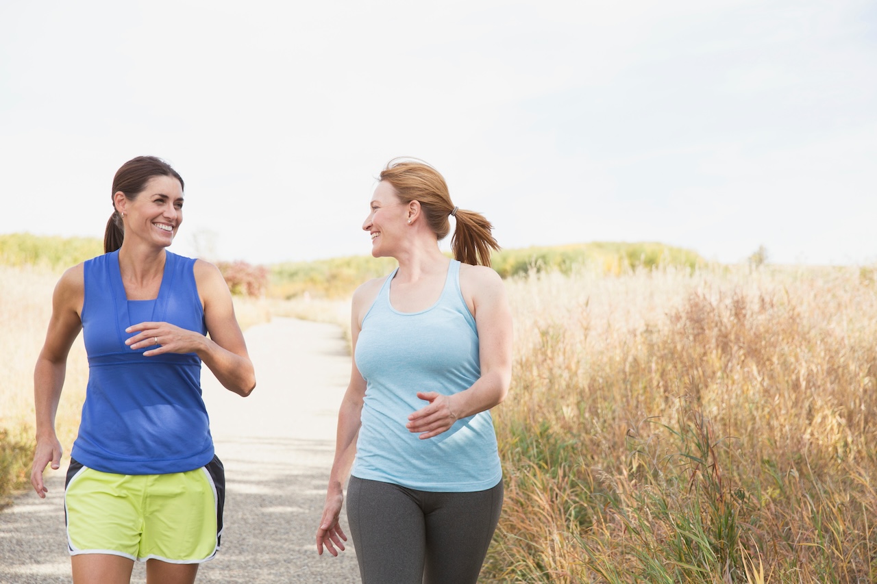 Two women jogging outdoors on a sunny day with tall grass on either side of the path, implying the benefits of walking as an exercise.