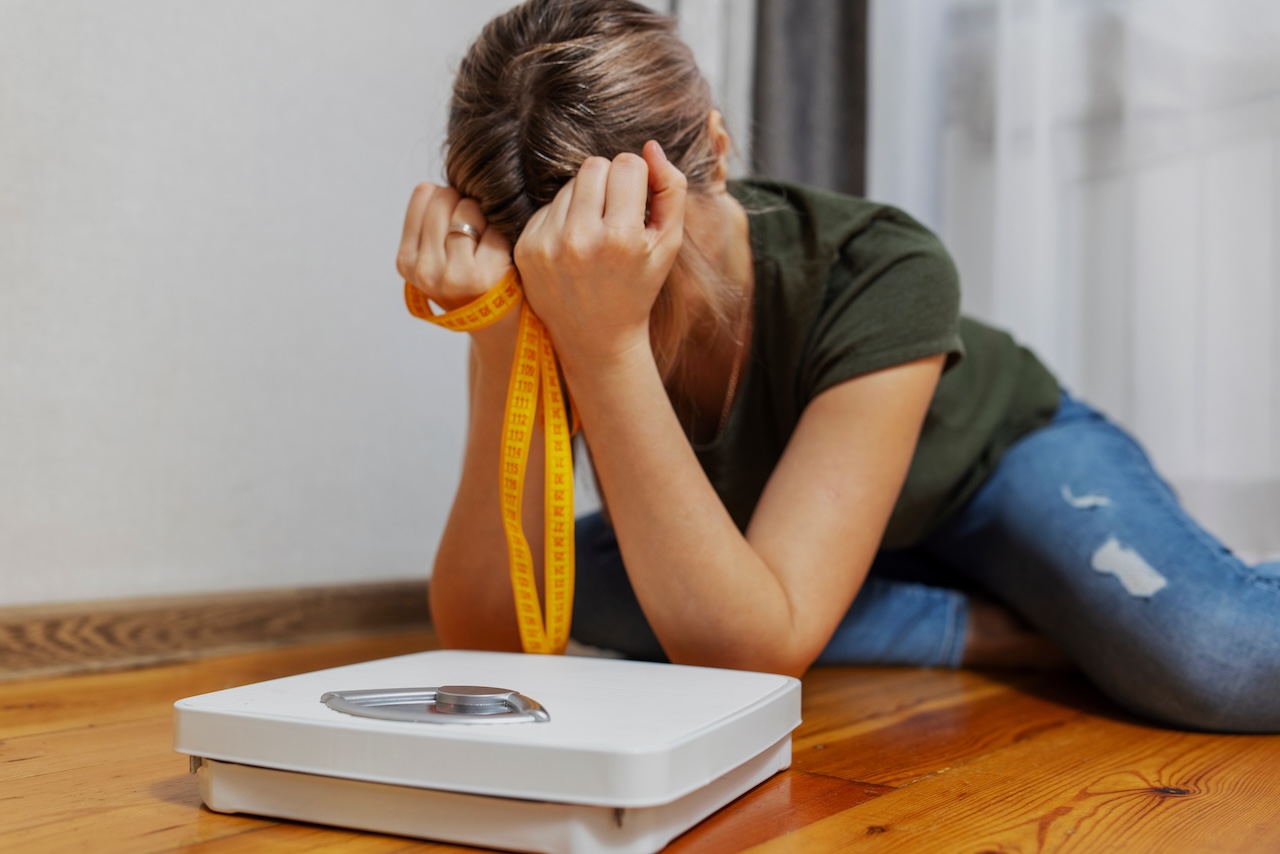 A woman sitting on the floor looking frustrated, holding a measuring tape around her neck, with a weight scale in front of her.