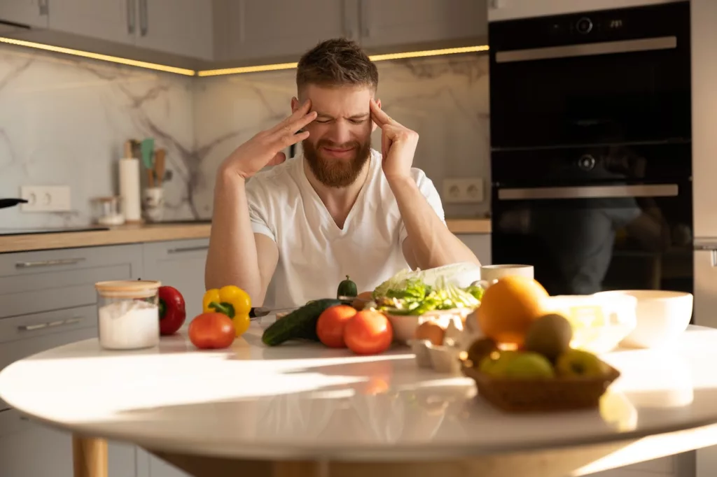 A man sits at a kitchen table, surrounded by fresh vegetables and fruits, pondering how many calories should I eat a day men.