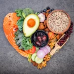 A heart-shaped arrangement of foods that help lower cholesterol with diet, including salmon, avocado, nuts, quinoa, and various fresh vegetables.