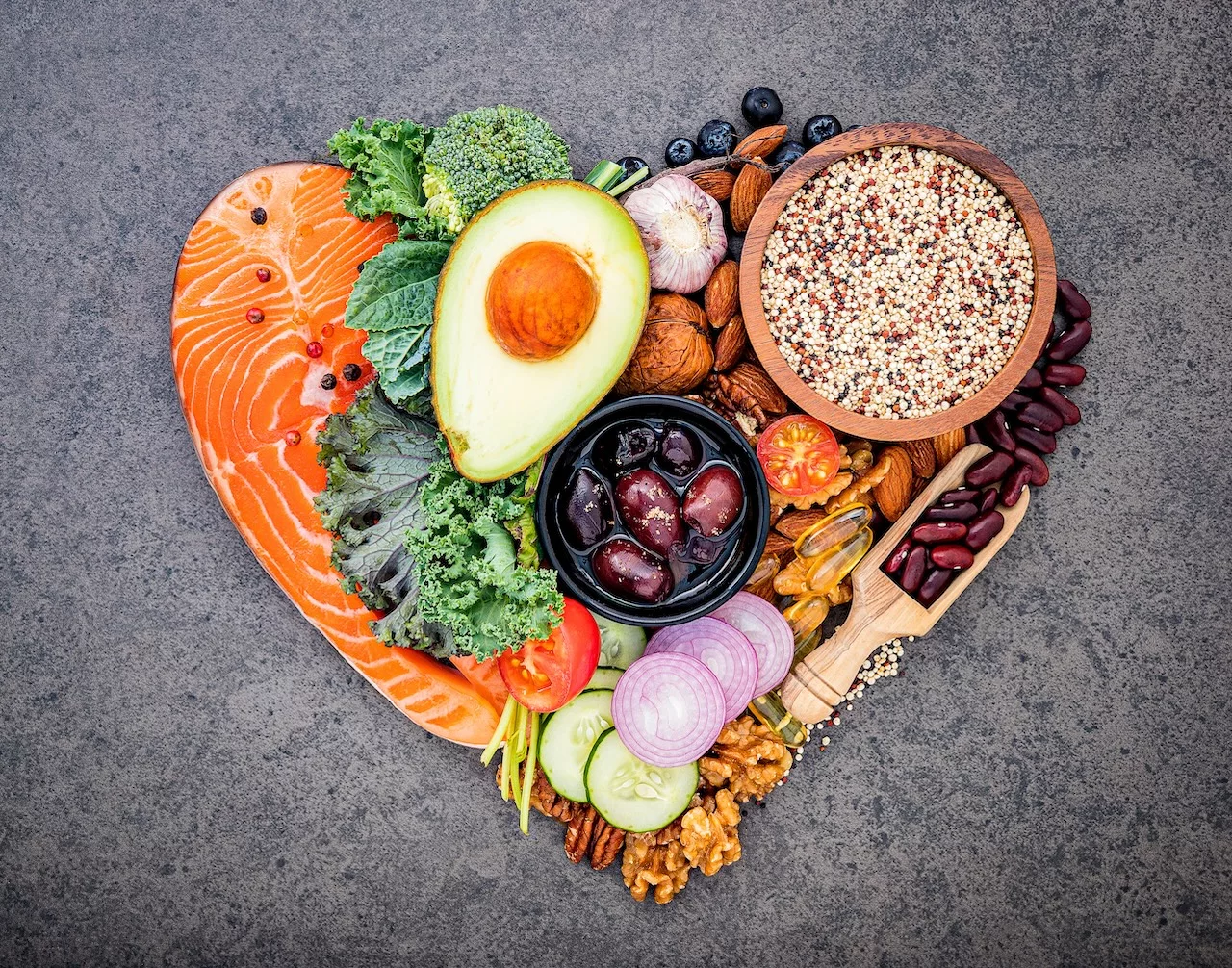 A heart-shaped arrangement of foods that help lower cholesterol with diet, including salmon, avocado, nuts, quinoa, and various fresh vegetables.